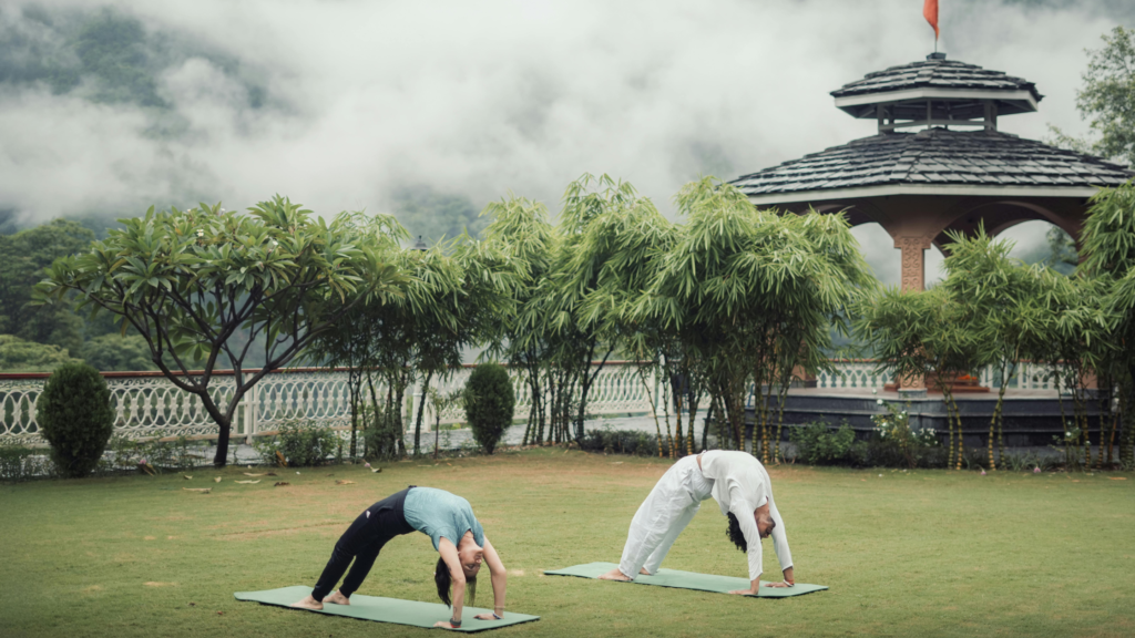 Two people practise yoga on a lawn as monsoon mist covers the hills behind, at a retreat in India